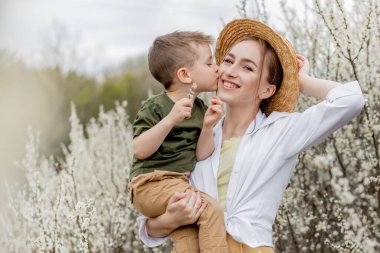 Mutlu anne ve oğul birlikte eğleniyor. Anne oğluna nazikçe sarılıyor. Arka planda beyaz çiçekler açar. Anneler Günü..