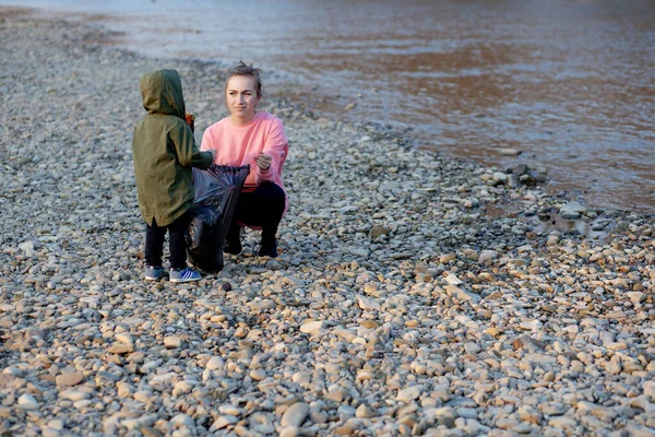 年轻妇女和她的儿子在河边的垃圾袋里收集塑料垃圾 空瓶使用的是肮脏的塑料瓶 海岸环境污染 — 图库照片