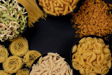 Tabaktaki farklı tipte kuru makarna ve siyah arka plandaki kaselerde. Metin için boşluk, üst görünüm.