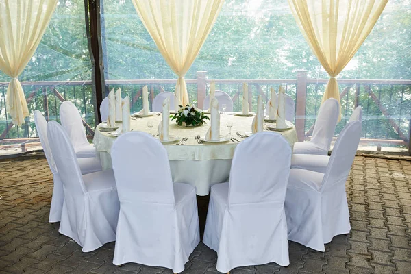 Wedding. Banquet. The chairs and roundtable for guests served with cutlery, flowers, and crockery and covered with a tablecloth