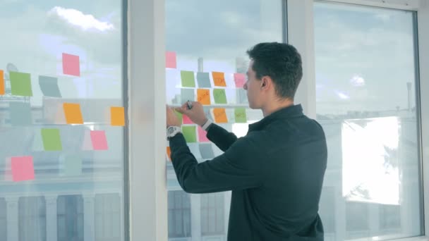 Lässiger junger Geschäftsmann Brainstorming-Schrift auf klebrigen Zetteln am Fenster im Büro. — Stockvideo