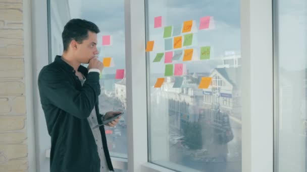 Casual νεαρός επιχειρηματίας brainstorming γραπτώς σε κολλώδεις σημειώσεις σχετικά με το παράθυρο στο γραφείο. — Αρχείο Βίντεο