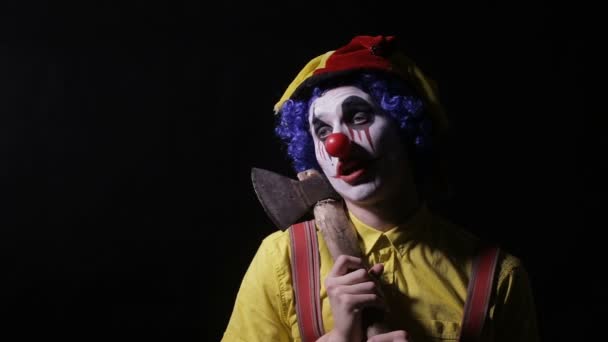 Enge clown met een bijl in een donkere kamer. Beangstigend nar, clown, Hansworst. — Stockvideo