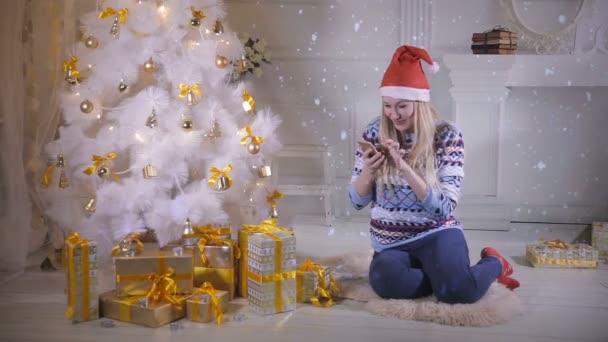 Pretty girl use mobile phone, sitting near Christmas tree during Christmas Eve. — Stock Video