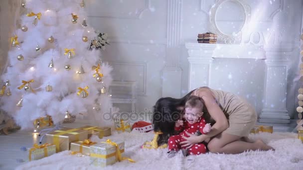 Mutter und Baby lachen und amüsieren sich in der Nähe des geschmückten Neujahrsbaums voller bunter glitzernder Lichter. — Stockvideo