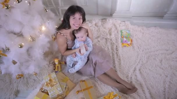 Weihnachtsfamilienkonzept. Mutter füttert ihr Baby während Neujahrsfeier in der Nähe des Weihnachtsbaums. — Stockvideo