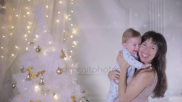 Madre y bebé riendo, divirtiéndose cerca del árbol de año nuevo decorado lleno de luces brillantes coloridas . — Vídeos de Stock