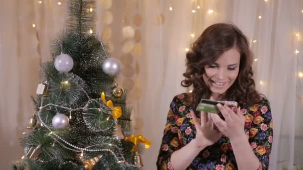 Joven linda chica emocional comprar regalos con tarjeta de crédito y teléfono inteligente en Navidad cerca del árbol de Navidad . — Vídeo de stock