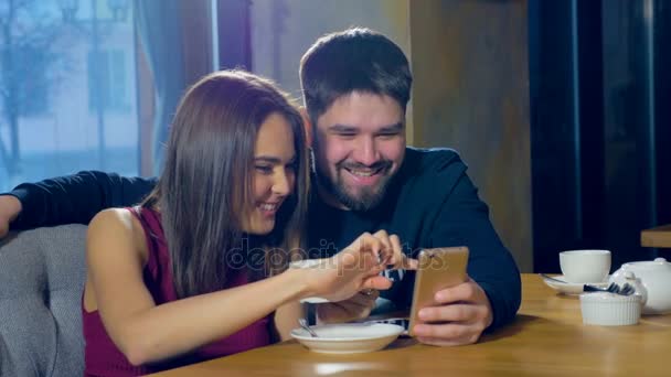 Joven hombre y mujer en la cafetería utilizando la nueva aplicación en el teléfono inteligente. Sonriendo feliz, adolescentes casuales . — Vídeo de stock