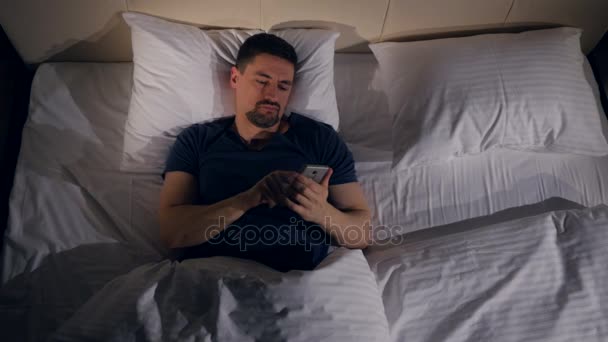 Счастливый смеющийся мужчина смотрит в телефон, лежа в кровати. 4K . — стоковое видео