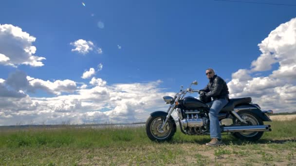 Un motociclista, un motociclista descansa en su motocicleta en un campo verde bajo las nubes. 4K . — Vídeo de stock