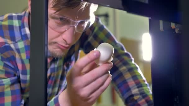 Un hombre sostiene una pequeña muestra de cabeza de muñeca y sonríe a una impresora 3D en funcionamiento. 4K . — Vídeo de stock