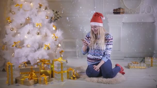 En ung kvinna kontrollerar sin smartphone under en julgran. — Stockvideo