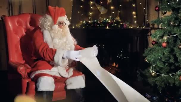 Papai Noel se senta em sua cadeira na frente de uma árvore de Natal lendo lista de presentes de ano novo . — Vídeo de Stock