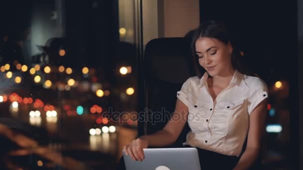 Uma mulher de negócios sorri e trabalha em um laptop perto de uma janela escura . — Vídeo de Stock