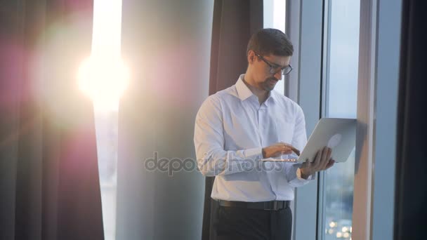 Un hombre de negocios se para cerca de una ventana y escribe en un portátil . — Vídeo de stock