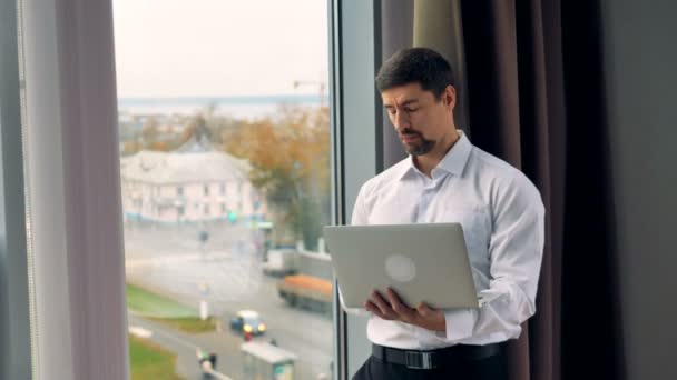 Un hombre de negocios trabaja en un portátil de plata de pie cerca de una ventana abierta durante el día . — Vídeo de stock