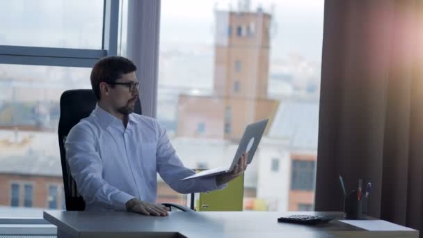 Un hombre de negocios se acerca a una ventana de la oficina para escribir en una computadora portátil . — Vídeos de Stock