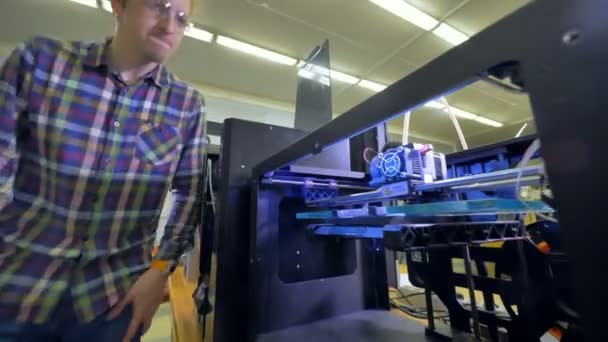 Un ingeniero se acerca a mirar por encima de una gran impresora 3D que comienza su trabajo. 4K . — Vídeo de stock