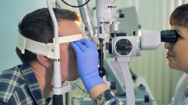 Ένα θηλυκό οφθαλμίατρο ελέγχει νέοι επανδρώνει τα μάτια με σχισμοειδή λυχνία σε μια κλινική. 4k. — Αρχείο Βίντεο