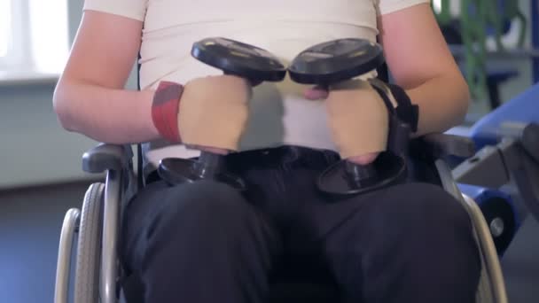 En vuxen man sitter i rullstol, håller två tyngdlyftningsvikter och tittar in i kameran — Stockvideo