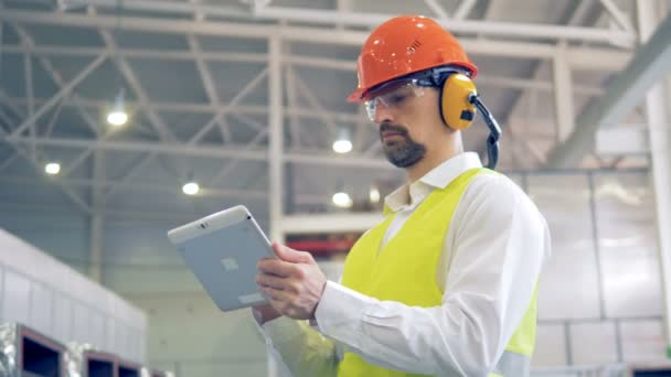 Un trabajador con sombrero duro y auriculares escribe información sobre su gadget, mirando las mercancías en un almacén . — Vídeos de Stock