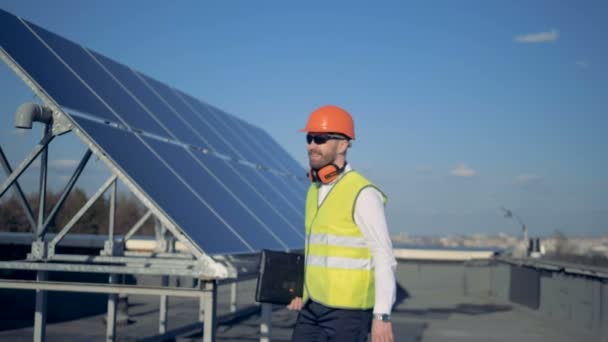 Um homem com um laptop sorri para a câmera, de pé perto de baterias solares no telhado. 4K . — Vídeo de Stock