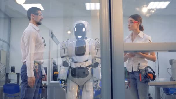 Labb arbetstagaren ger riktningar för en robot som genomförs av en kvinnlig ingenjör med en dator — Stockvideo