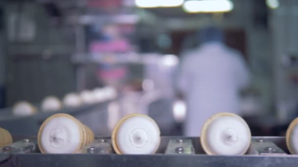 Primer plano de las copas de gofres con helado moviéndose a lo largo del transportador con los trabajadores de la fábrica en el fondo — Vídeos de Stock