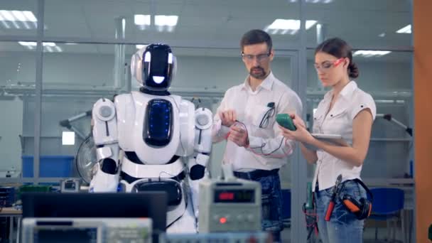 Male and female engineers are taking readings of a human-like robot with special equipment — Stock Video