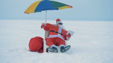 Santa Claus is sitting in a deck-chair with a tablet and his umbrella is getting blown away