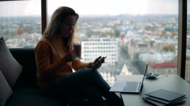Bir kadın kahve ve telefonla şehir manzarasının yanında oturuyor.
