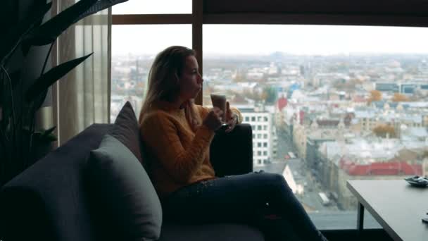 Жінка п'є каву і дивиться у вікно з видом на місто — стокове відео