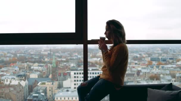 Jovem mulher está sentada contra uma vista da cidade e beber café — Vídeo de Stock