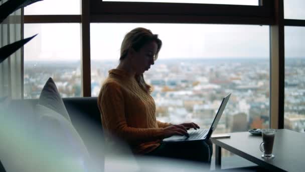Una señora está escribiendo en un portátil en un café con una vista urbana — Vídeo de stock