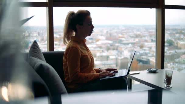 Uma mulher com um computador está trabalhando em um café com vista para a cidade — Vídeo de Stock