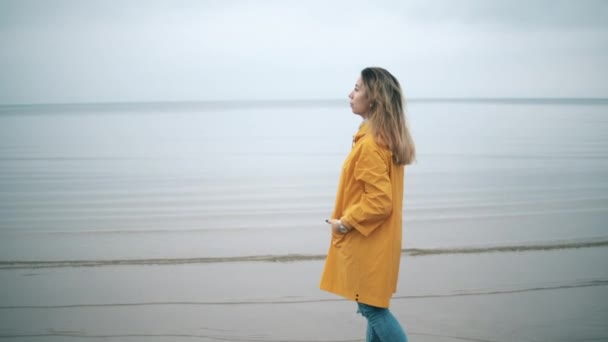 Una mujer con un abrigo amarillo está de pie en la costa — Vídeo de stock