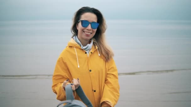 Señora sonriente en un abrigo amarillo está usando gafas de sol cerca del mar y el cielo nublado en el fondo . — Vídeo de stock