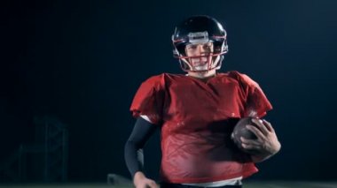 American football player walks on a field, holding a ball in hands, close up.