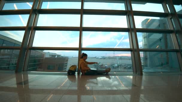 Жінка друкує на ноутбуці, сидячи на підлозі в аеропорту — стокове відео