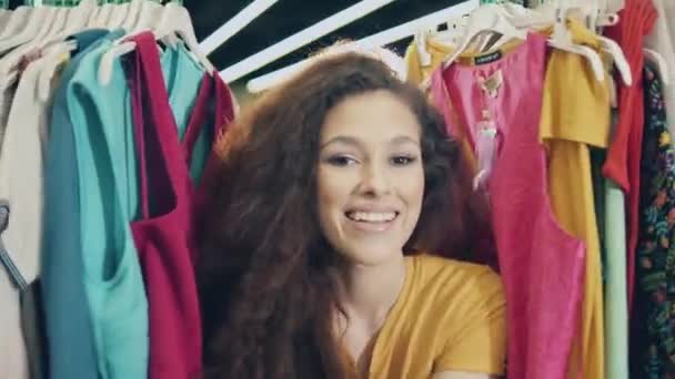 Mujer joven está sonriendo entre los vestidos en una vista de retrato. Feliz concepto de compras. — Vídeos de Stock