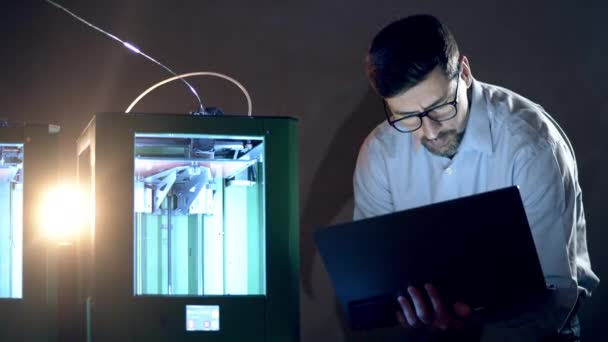 Een man met laptop bestuurt een 3D printer. 3d drukindustrie research engineer werkzaam in het lab. — Stockvideo