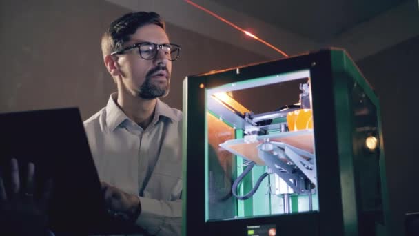 Çalışan adam 3D yazıcıyı dizüstü bilgisayarla kontrol ediyor. Bir tasarım stüdyosunda 3D yazıcıyla çalışan bir mühendis.. — Stok video