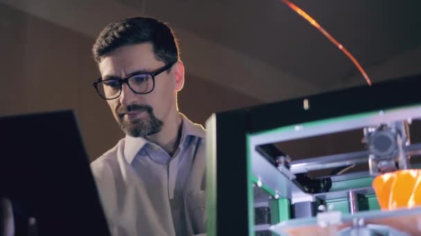 Un ingeniero con un portátil está viendo la impresora 3D en el trabajo — Vídeo de stock