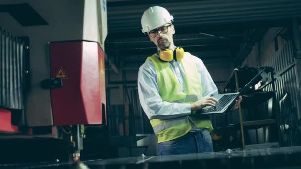 Industriële werknemer typt op een laptop terwijl hij naar de lasersnijder kijkt — Stockvideo