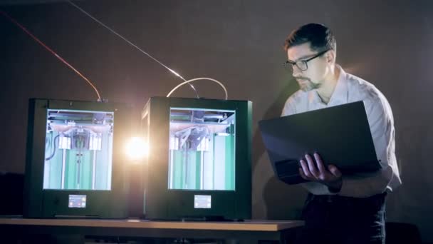 Mannelijke ingenieur bestuurt 3D-printers via zijn laptop. Man werkt met een 3D printer bij 3d printing lab. — Stockvideo