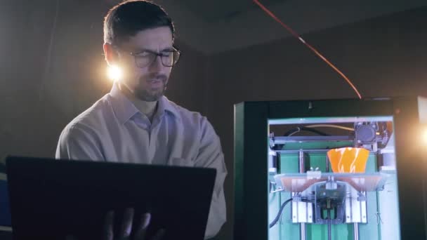 Un técnico con una computadora portátil está examinando una impresora 3D en funcionamiento — Vídeo de stock