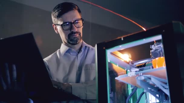 Ein Ingenieur bedient einen Laptop, während er einen 3D-Drucker anschaut. 3D-Drucker, der einen Gegenstand aus Kunststoff druckt — Stockvideo