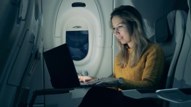 Καμπίνα το βράδυ με μια κυρία να χαμογελάει ενώ πληκτρολογεί σε ένα φορητό υπολογιστή. Γυναίκα ελεύθερη επαγγελματίας που εργάζεται σε φορητό υπολογιστή κάθεται στο αεροπλάνο. — Αρχείο Βίντεο