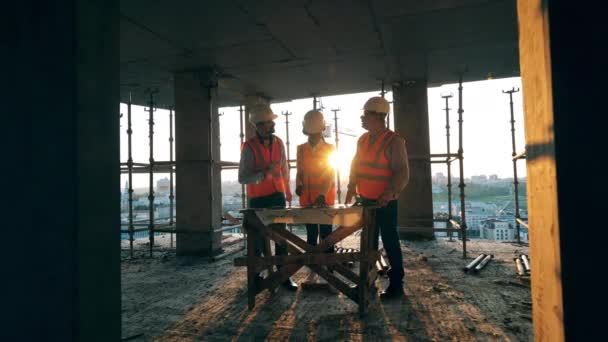 Les travailleurs parlent dans un bâtiment inachevé sur un fond de coucher de soleil. Ingénieurs multiethniques, architectes discutant du plan de construction . — Video